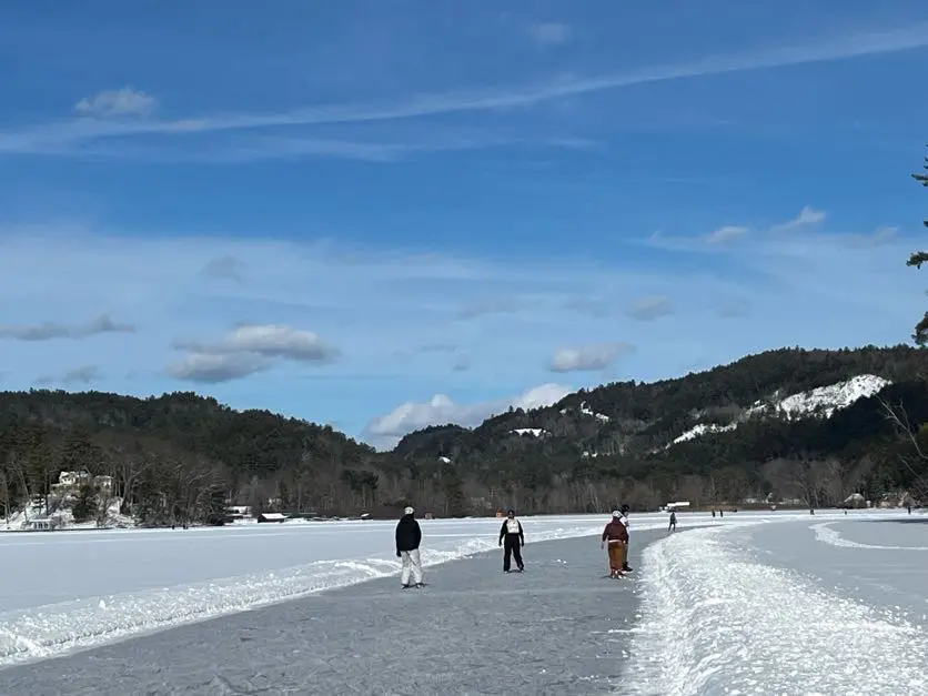A group of 3 people skate on a ice trail on a snow-covered lake under a blue sky in front of snowy hills.