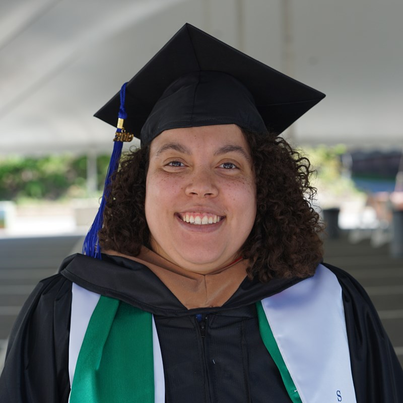 Headshot of Jeanine Buonopane in a cap in gown outside of the Tsongas Center at UMass Lowell at Commencement 2019