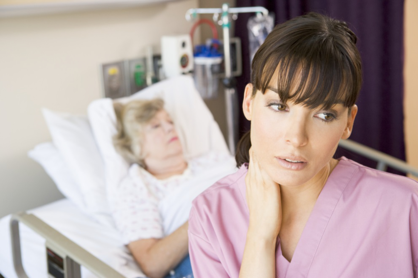 Image of a nurse in foreground looking stressed with an older woman patient lying in a hospital bed behind her. The image depicts the need for the job stress program which would help nurses learn how to minimize stress, which would in turn translate to improved patient care.