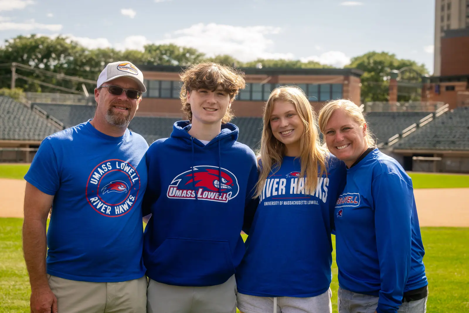 Father, son, daughter and mother wearing UMass Lowell River Hawks clothing standing on ball field