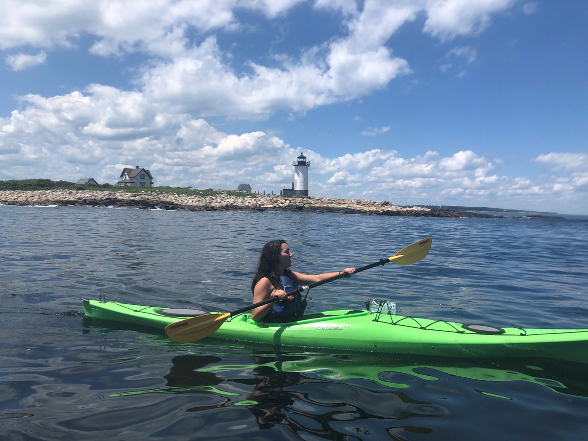 one woman paddles a solo kayak on the ocean with a lighthouse behind her