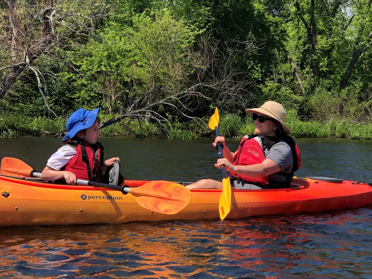 A mom and her child paddle a kayak with green shore behind them