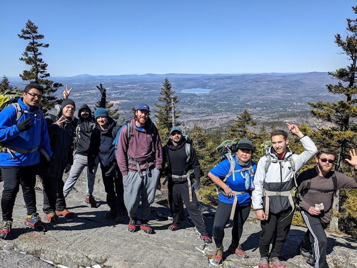 A group of 9 people smile at the top of an overlook with mountains and a pond beyond