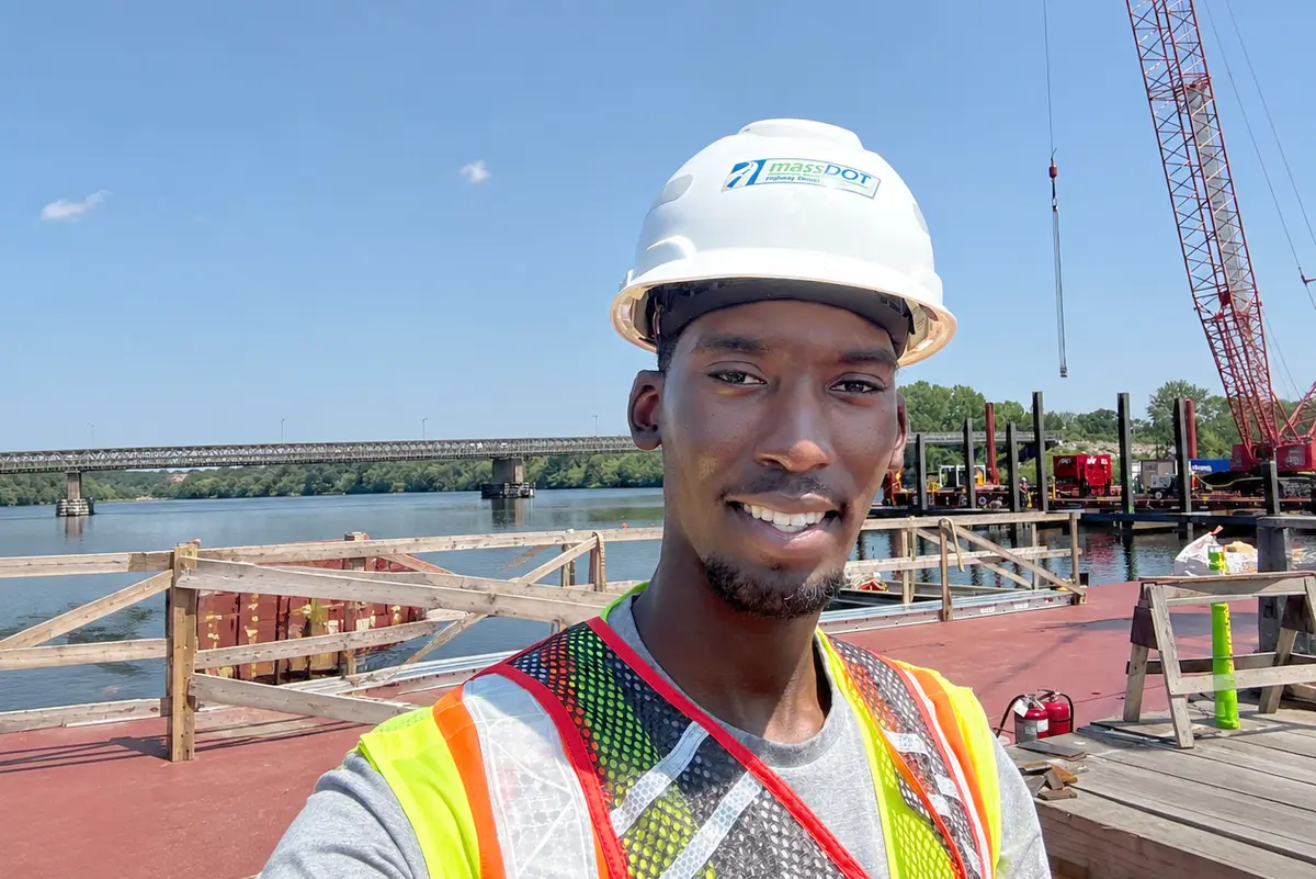 A young man in a hard hat and safety vest poses for a photo standing on a construction barge on a river with a bridge in the background.