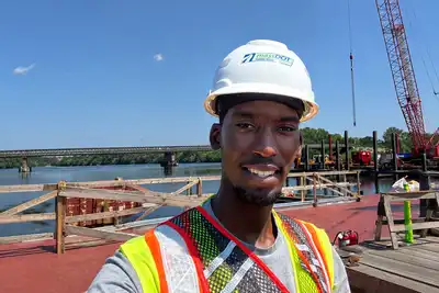 A young man in a hard hat and safety vest poses for a photo standing on a construction barge on a river with a bridge in the background.