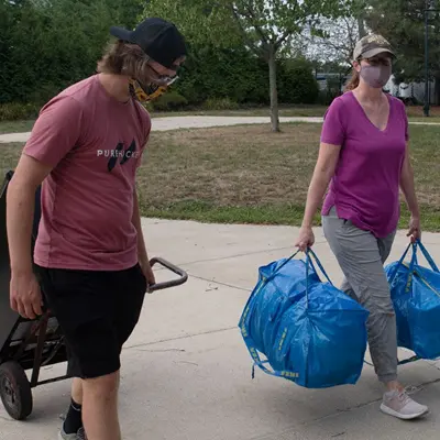two students wearing masks and baseball caps carrying luggage