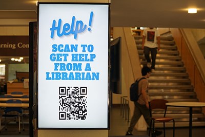 Students walk near a staircase behind a monitor with a QR code for library help