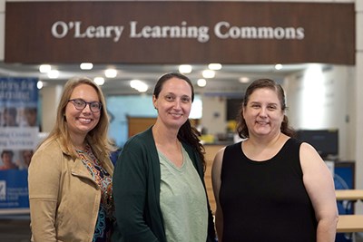 Three women smile while posing for a picture in front of a sign that says O'Leary Learning Commons