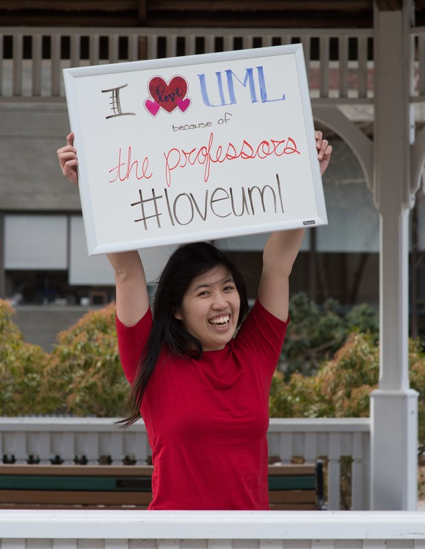 Female UMass Lowell student smiling and holding up a whiteboard that reads "I love UML because of the professors #loveuml"