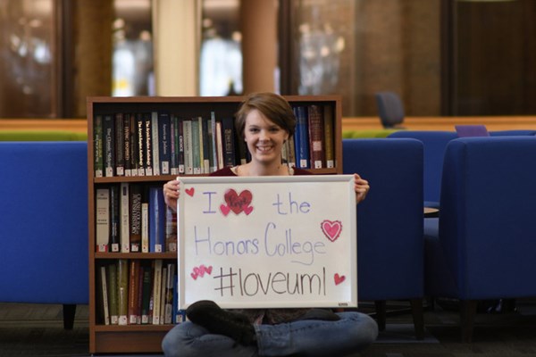 An honors student sitting in O'Leary Library holding a whiteboard that says "I love the Honors College #loveuml" with hearts on it