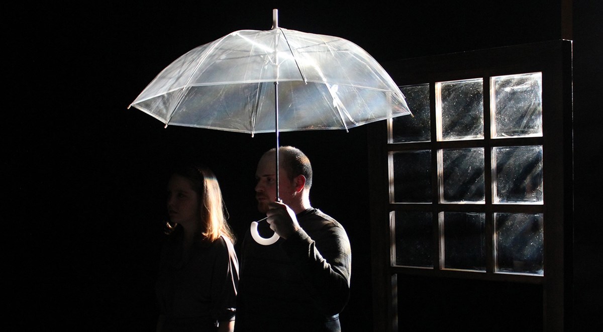 A male student holding an umbrella on stage next a female student with an window behind them in the dark during a UMass Lowell Theatre Arts production.