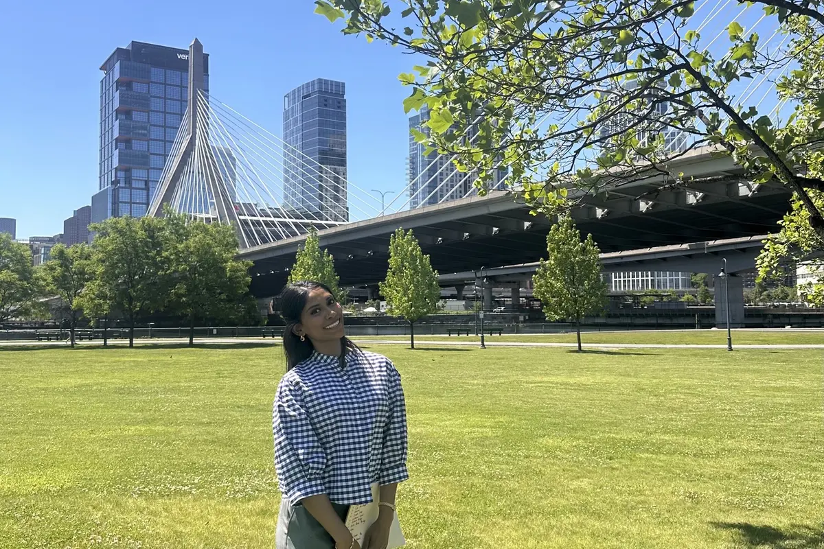 A young woman poses for a photo in a park with a bridge and buildings behind her.