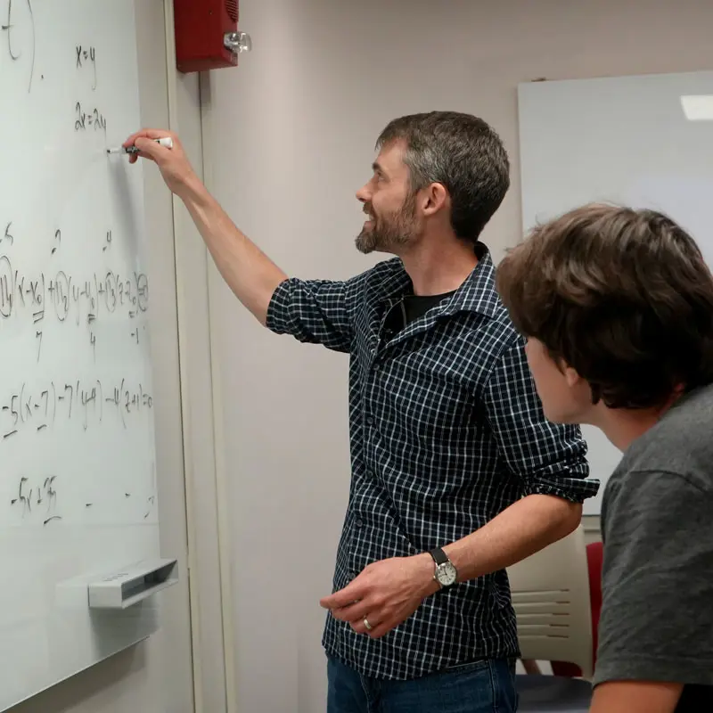 Professor writes an equation on a whiteboard while a student watches.