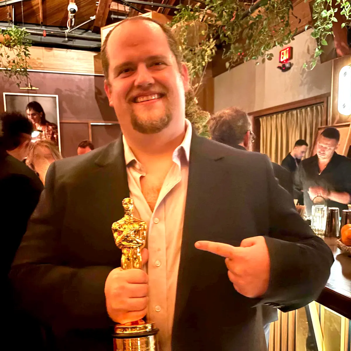 A man in a suit jacket smiles and points to an Oscar trophy while posing for a photo.