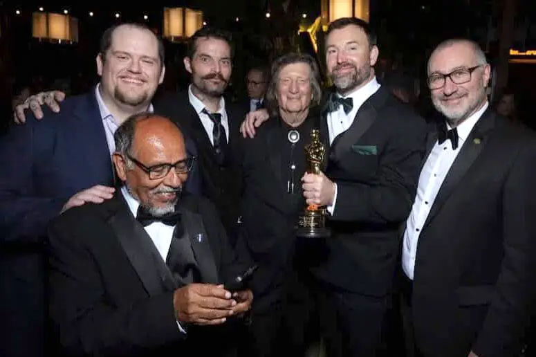 Six men in formal wear pose for a photo while one of them holds an Oscar trophy.