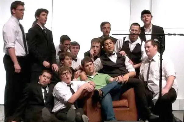 A dozen male college students perform on stage, with one of them sitting in a chair and rest gather around him.
