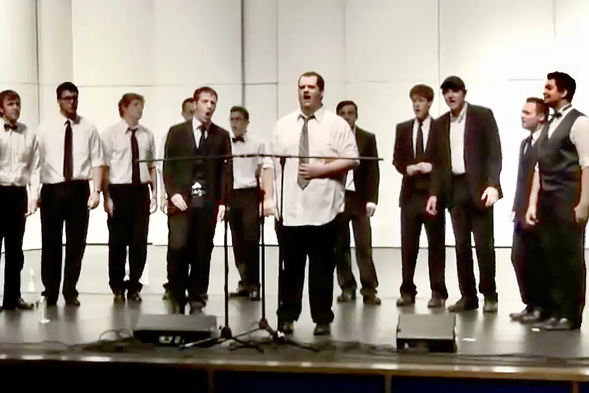 A young man sings on stage with a dozen others singing behind him.