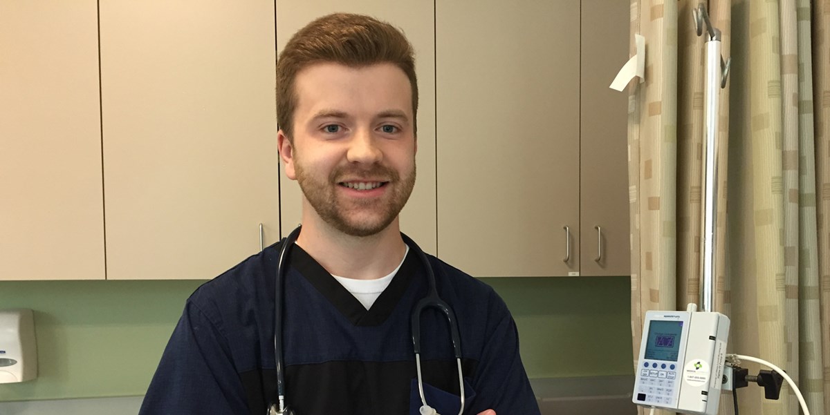 Matthew Fowler wearing scrubs and a stethoscope in a nursing simulation lab at UMass Lowell