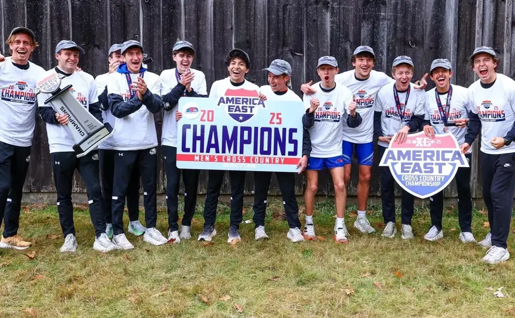 Members of the UMass Lowell men's cross country team lined up in front of a fence holding America East 2025 Men's Cross Country Champions banners