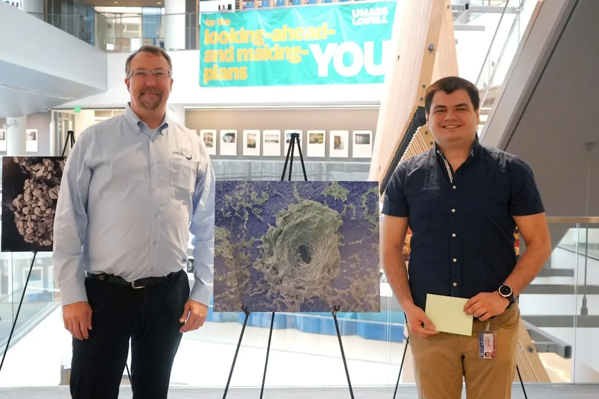 Two men stand before winning photo in front of banner that says for the looking ahead and making plans you UMass Lowell.