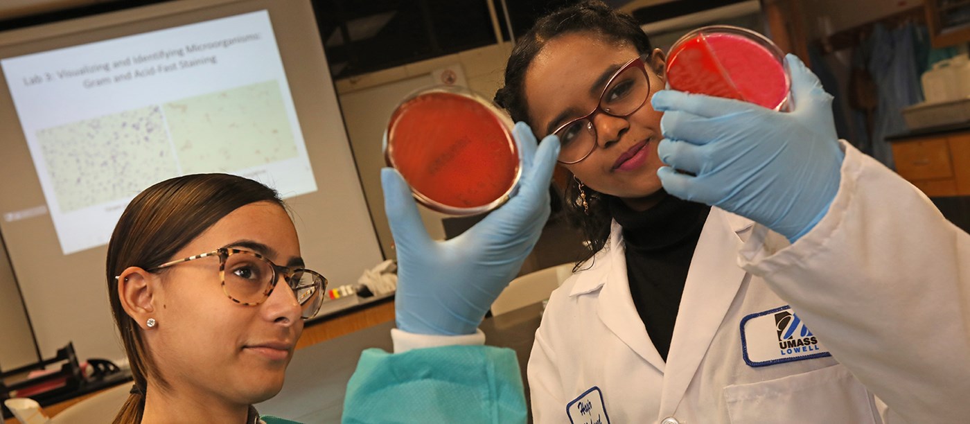 Two female students wearing lab gear look at dishes of red matter