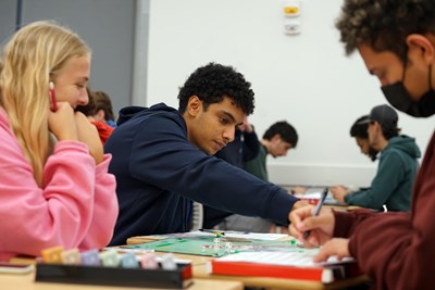 A young man moves a piece on a Monopoly board while another player in a pink sweatshirt looks on