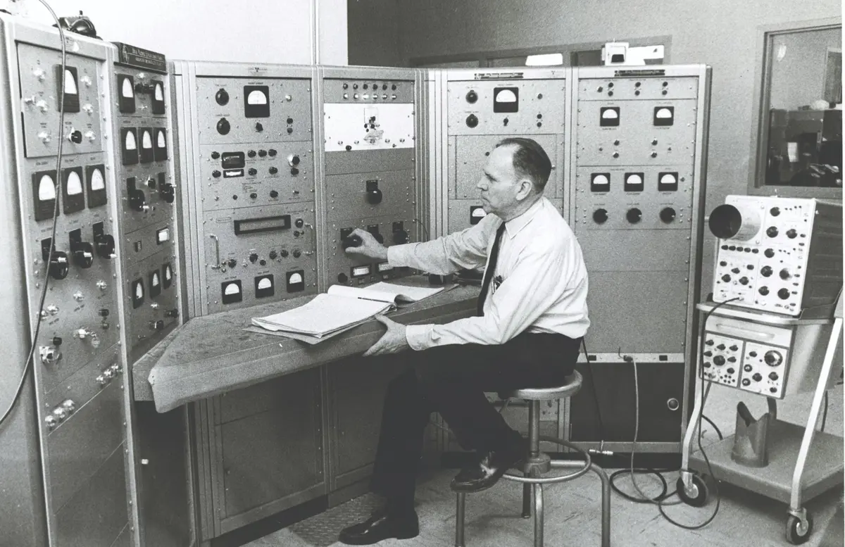 Man sits in front of wall of dials and gauges in lab in black and white photo