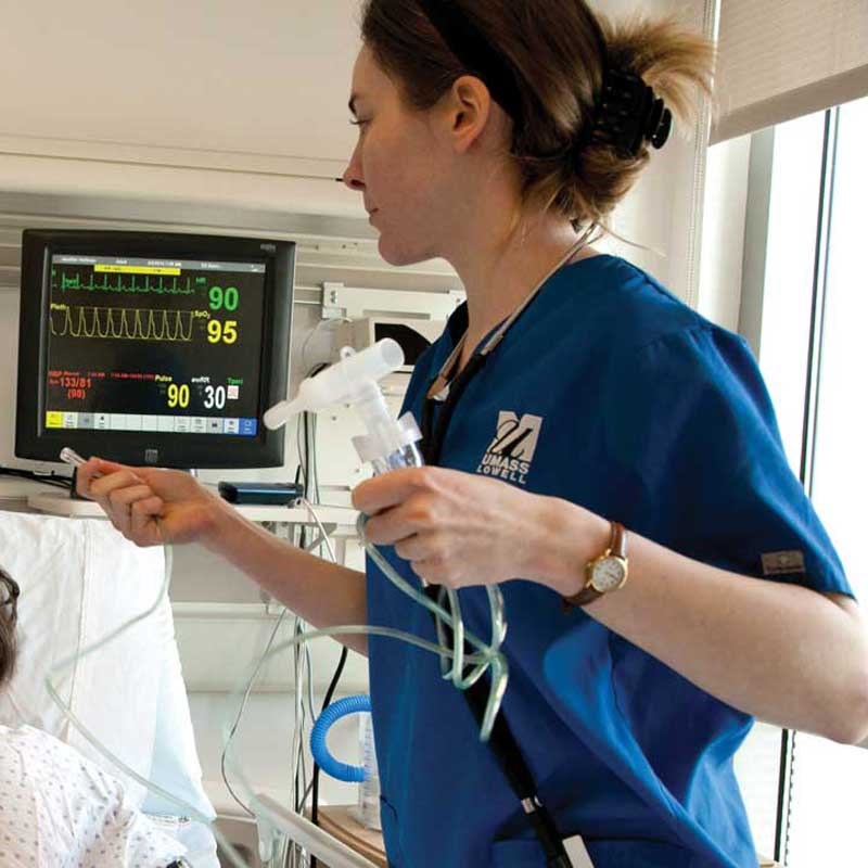 Nursing student wearing blue scrubs holds tubes near a patient bed in a UMass Lowell demonstration hospital wing