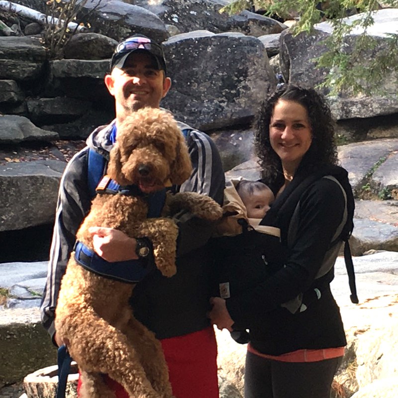 UMass Lowell alumni John and Ella pose in front of a Rocky landscape with their six-month-old son and Golden Doodle puppy
