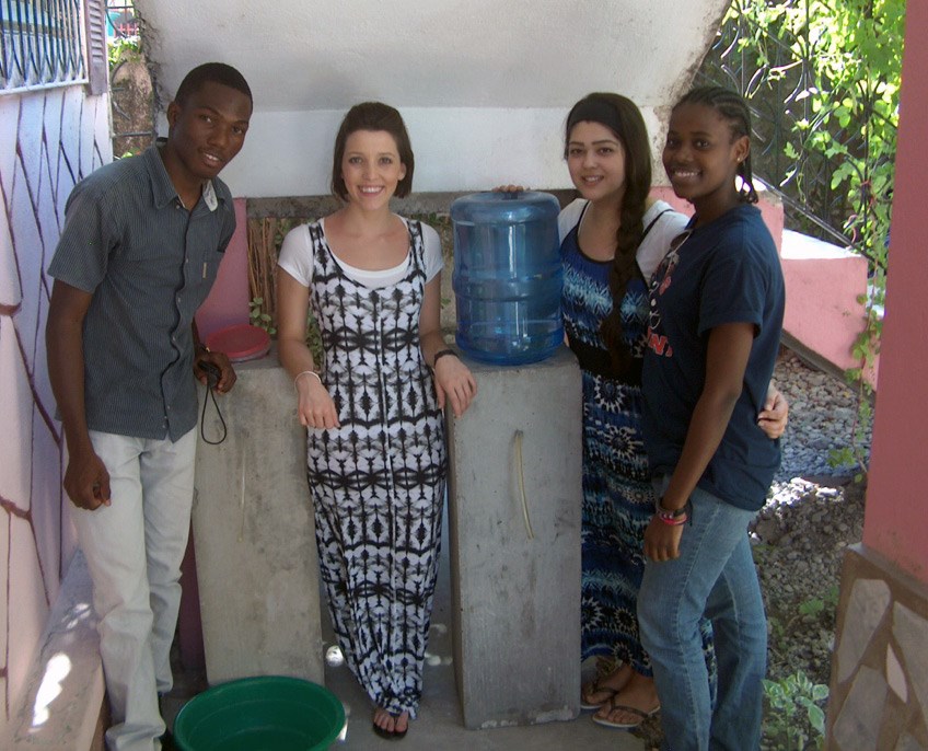 One male young adult and three female young adults posing for a picture next to a water jug with a hose for dispensing.