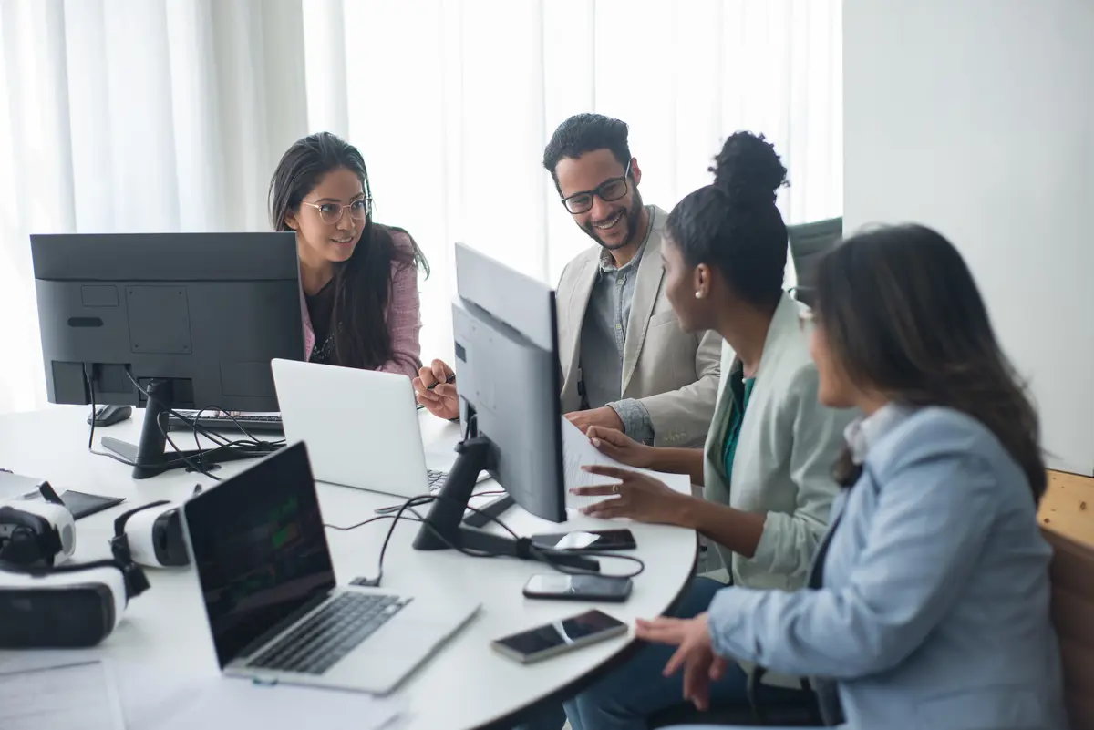4 people talking and smiling at a table with multiple computers and phones in an office.