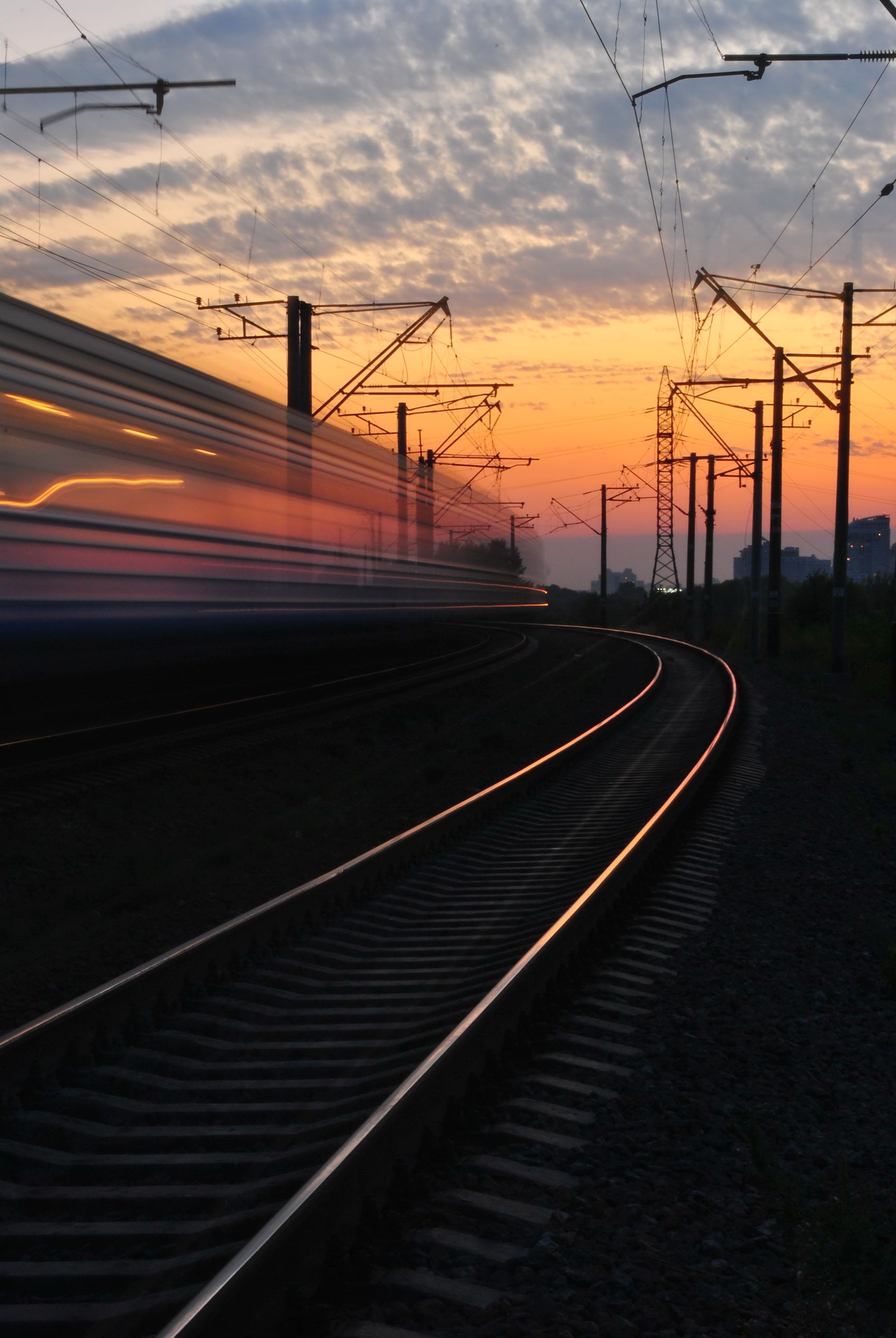 A train blurs at high speed against a sunset in front of electrical towers