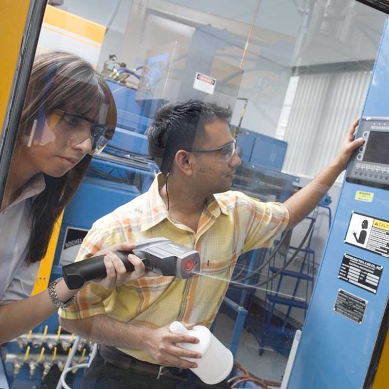 Two UMass Lowell plastics engineering students seen through window of a Blow Molding machine