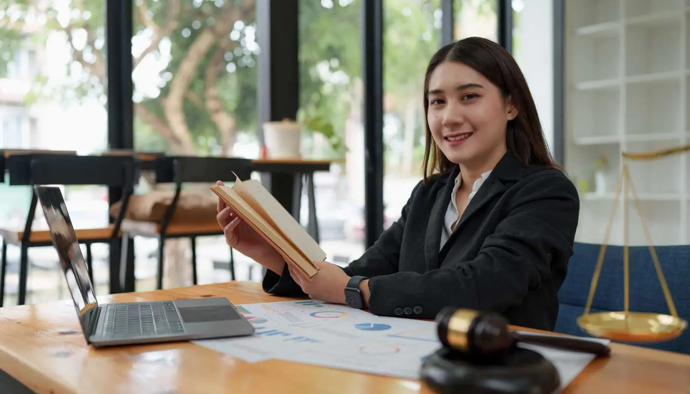 Student holds a book while seated at a desk with a laptop and judicial gavel.