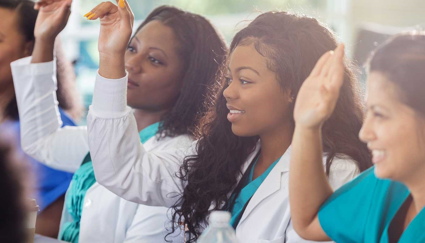 Three premed students wearing medical coats raise their hands in a classroom. 