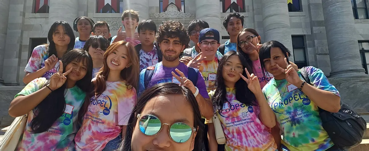 Sixteen high school students in tie-dye t-shirts pose for a group selfie in front of a building.