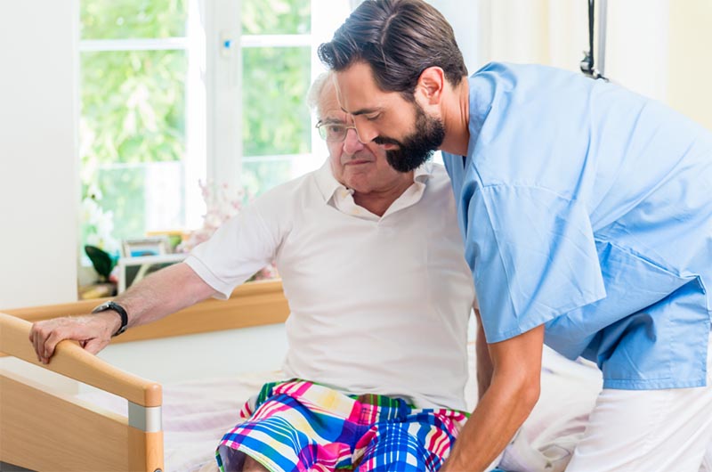 Picture of a male nurse helping an older man get out of bed efficiently and without injury to either of them. It demonstrates the ergonomics required for safe patient handling