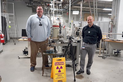 Two men stand next to research equipment and a radiation area caution sign