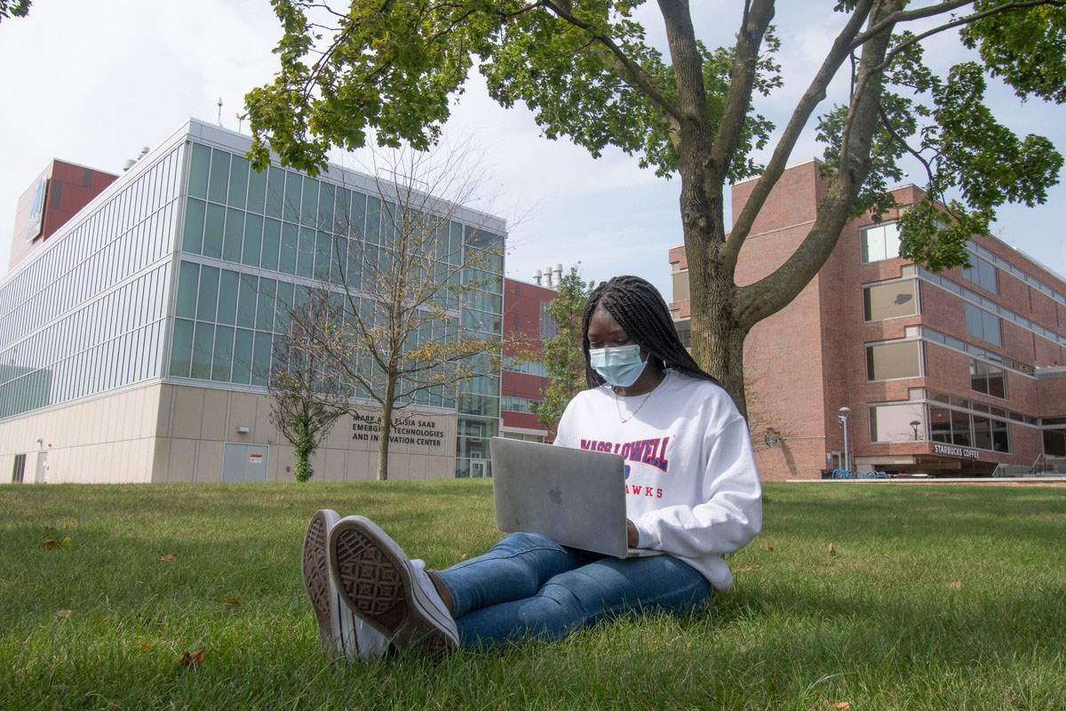 Young black woman wearing mask, seated on grass, working on laptop on her lap