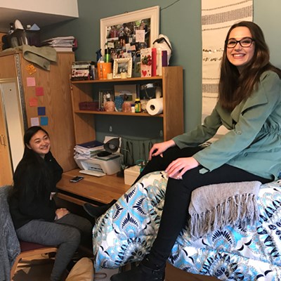 One female student sits at her desk in her room in her residence hall and her roommate sits on a bed, smiling at the camera