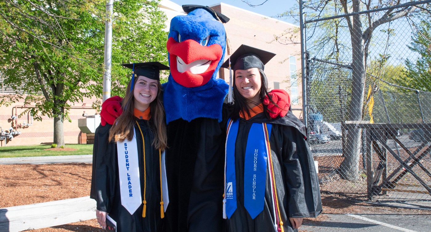 Two female graduates with Rowdy the River Hawk outside at Commencement