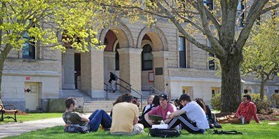 Group of students sit on the lawn in circle with laptops in front of Coburn Hall