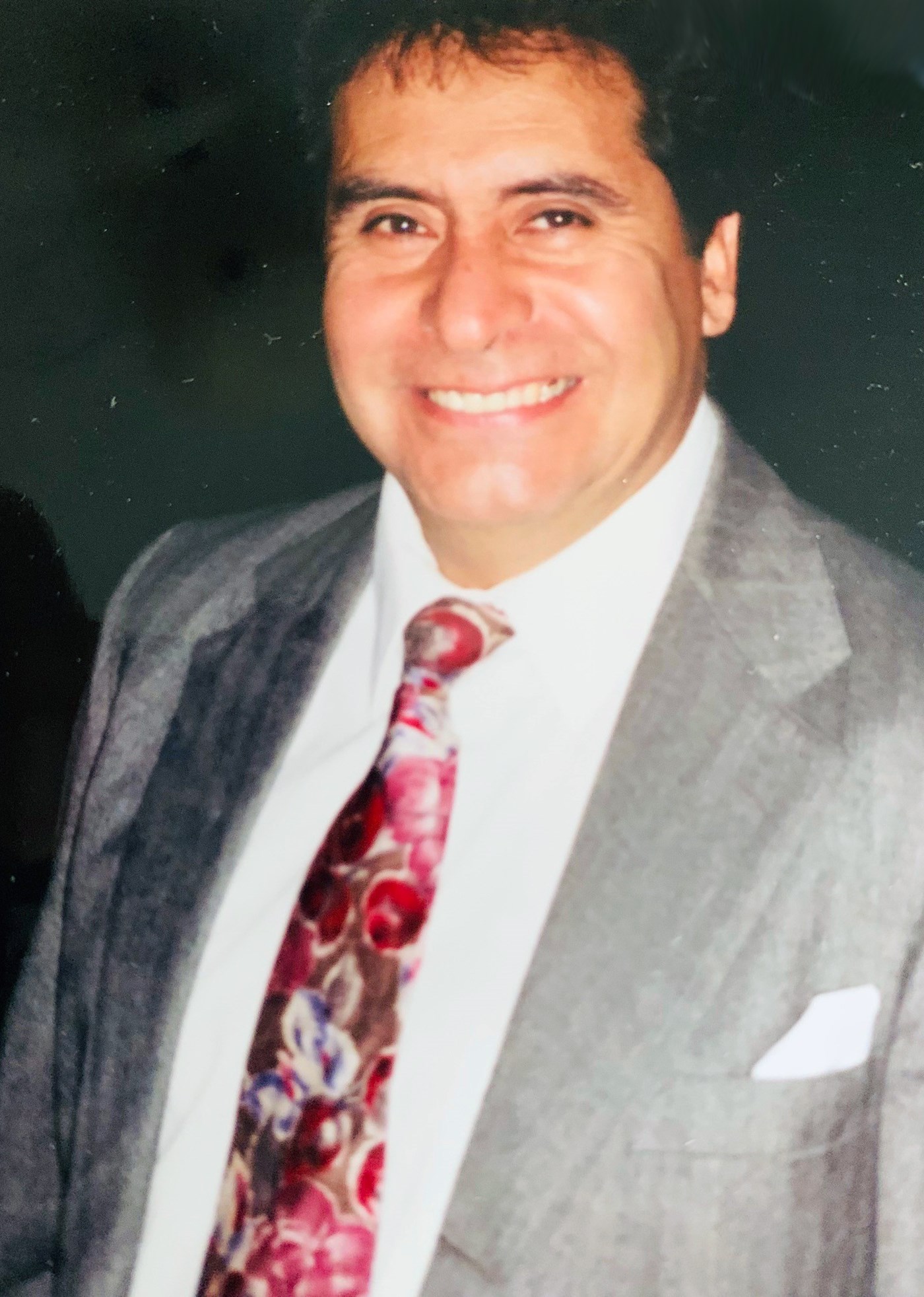 Juan Rodriguez wearing a grey suit, white short and red tie smiling at the camera.