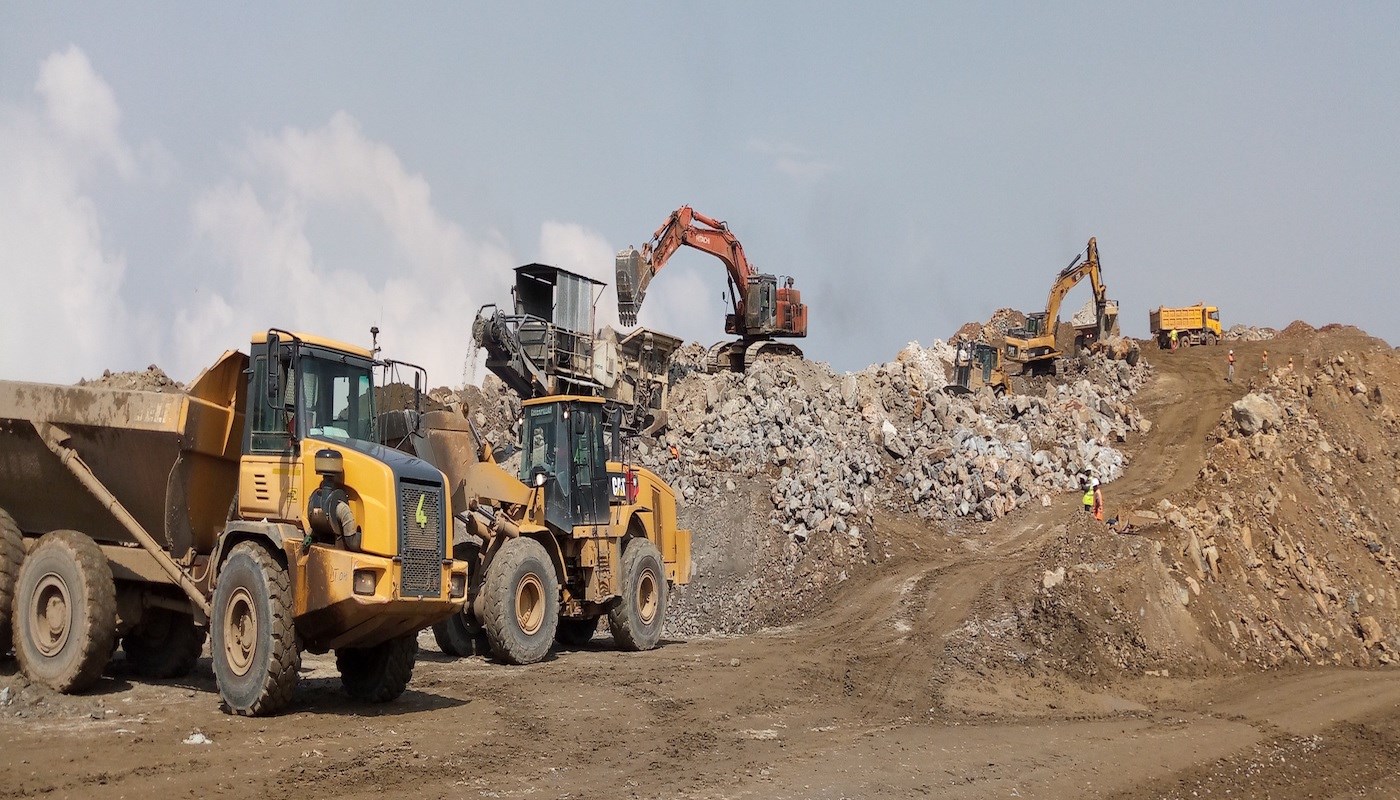 An average worksite for sand, stone, and gravel workers on a sunny day, without a cloud in the sky, containing large dust clouds in the background, and mining equipment, such as large flatbed trucks and excavators, surrounded my piles of sand and stone.