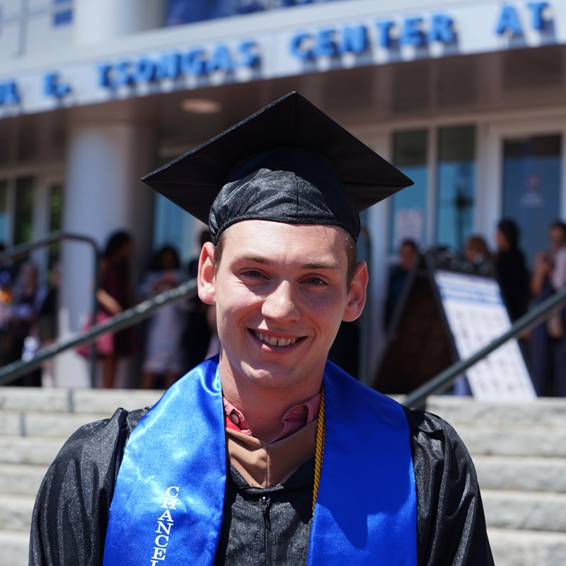 Headshot of Sean Flaherty in a cap in gown outside of the Tsongas Center at UMass Lowell at Commencement 2019