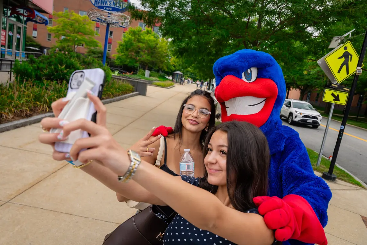 2 students pose outdoors on Move In Day for a selfie with mascot, Rowdy the River Hawk.