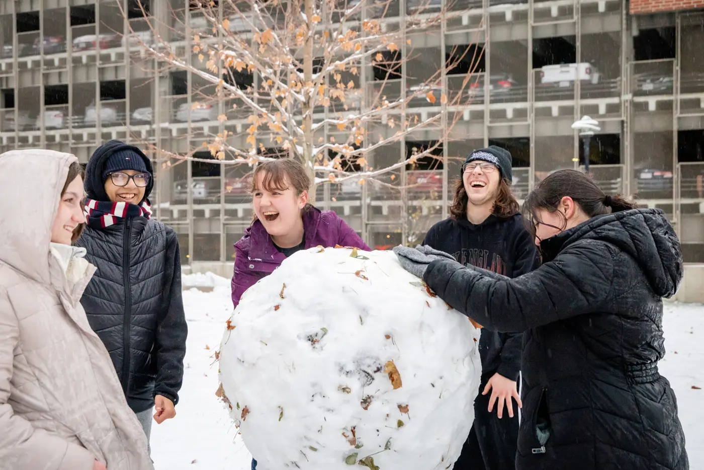Five laughing students in winter gear make a giant snowball in front of a building