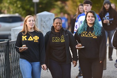 Three young women with sorority sweatshirts walk with plates of food
