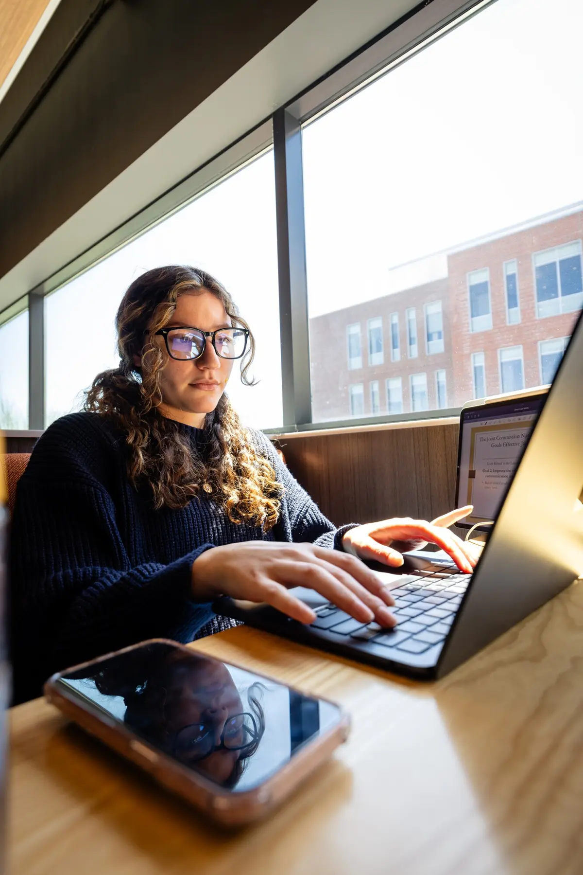Student wearing glasses typing on laptop with phone on desk and buildings outside window
