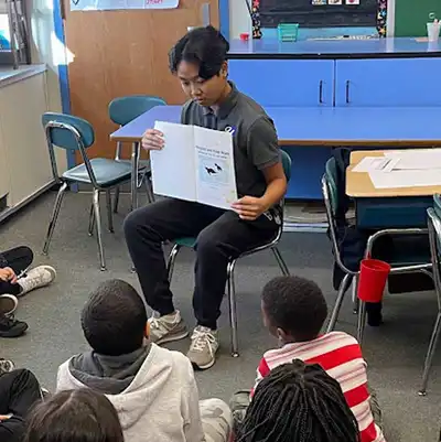A UMass Lowell student-teacher is holding a book and reading to elementary school children.